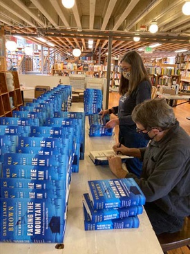 stacks of the hardcover of "Facing the Mountain" with Daniel James Brown (seated, masked) signing as Karen Maeda Allman (standing, masked) flaps copies for him inside Elliott Bay Book Company in Seattle. 
