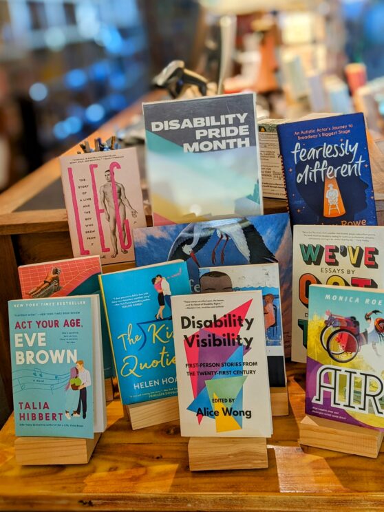 A display of books with a Disability Pride Month sign. Titles include Air, Fearlessly Different, We've Got This, Disability Visibility, Leg, The Kiss Quotient, Feathers Together, Act Your Age Eve Brown, Just BY Looking at Him, and Romance in Marseille