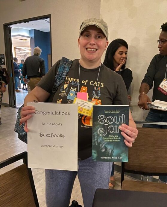 Photo of Annie Carl, a smiling woman in a t-shirt and baseball cap, holding sign announcing winner of Buzz Books t o be "Soul Jar" and a copy of the book, Soul Jar.