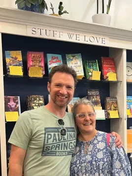 Brian in Paulina Books t-shirt and Sylla smile and pose in from of "Stuff We Love" bookshelf